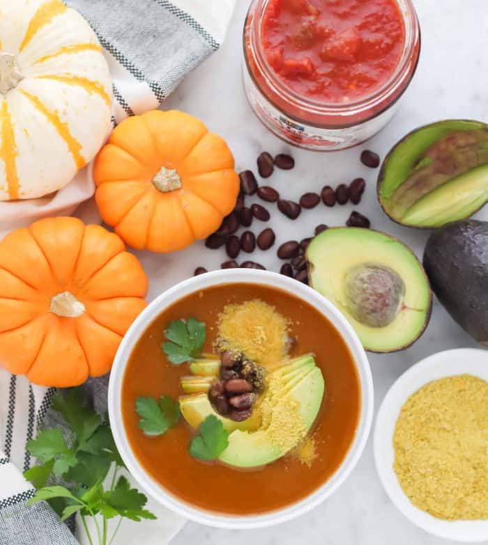 bowl of pumpkin soup topped with cilantro, avocado, and nutritional yeast with orange and white pumpkins on the side, salsa, avocado, nutritional yeast, black beans