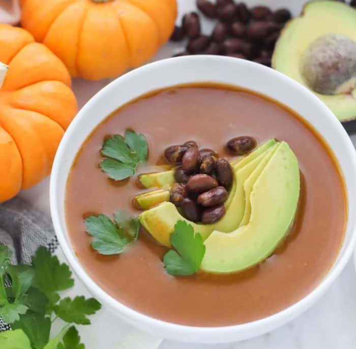 bowl of pumpkin soup topped with sliced avocado, black beans and cilantro, small pumpkins