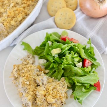 casserole dish and plate of poppy seed chicken casserole with green salad, on dish towel with crackers and onion