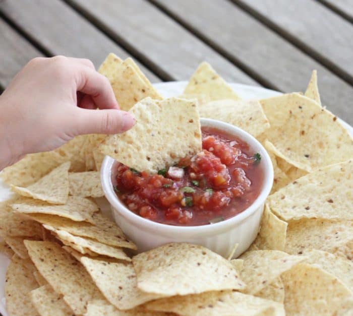 hand dipping a chip into Salsa in a bowl with a plate of chips