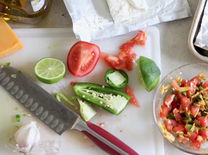 knife on a cutting board with cream cheese, cut jalapenos, tomatoes, lime