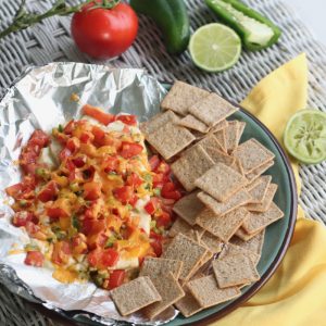 glass plate with foil topped with cream cheese appetizer and crackers on wicker table, yellow napkin and tomatoes, jalapenos and lime in background