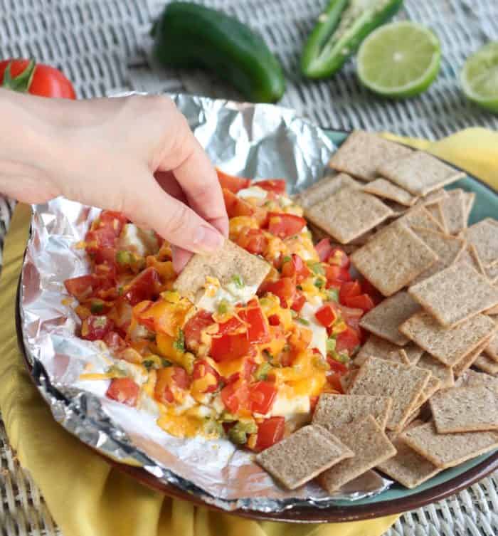 hand holding a cracker dipping into a cream cheese appetizer covered in tomatoes and cheese