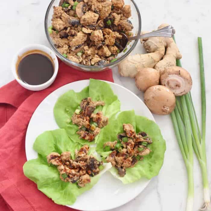 white plate with lettuce wraps on a red napkin with a bowl of sauce and turkey mixture with mushrooms and green onions