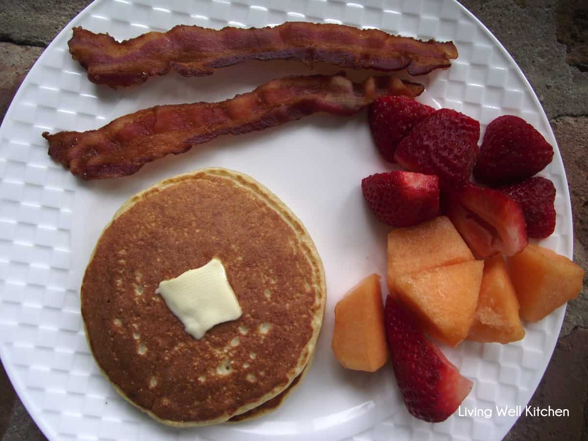 white plate with protein pancakes topped with butter, sliced strawberries and cantaloupe, and bacon.
