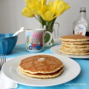 two pancakes topped with butter on a plate next to a stack of pancakes, flowers, a blue bowl, and a cup of tea.