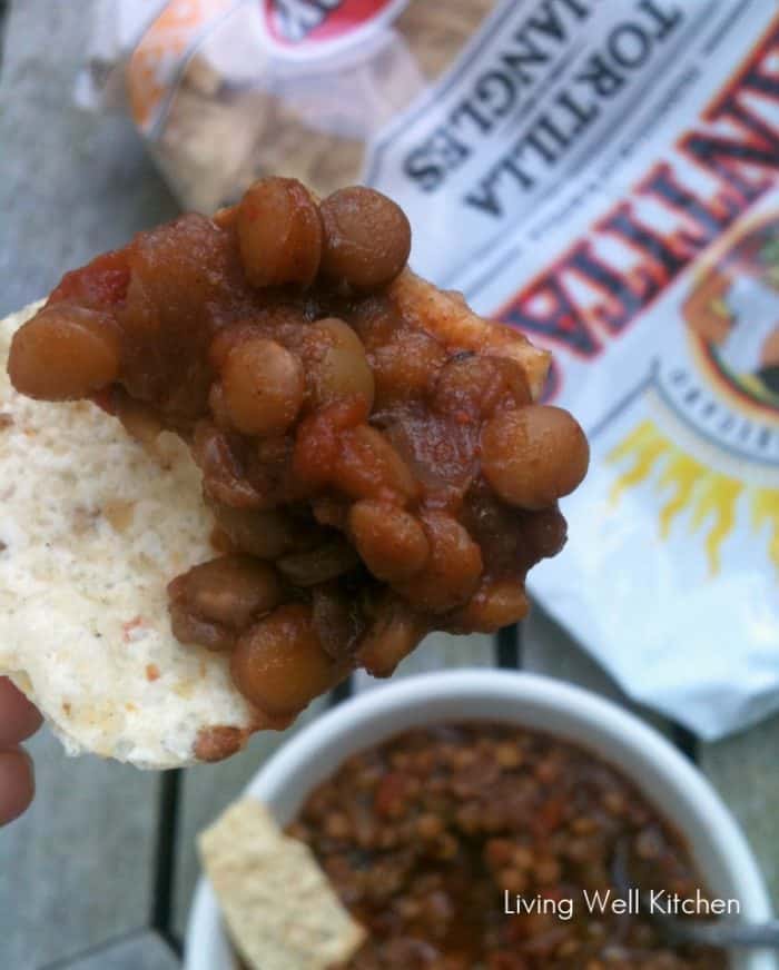 tortilla chip with lentil chili on top with a bag of chips and a bowl of soup in background