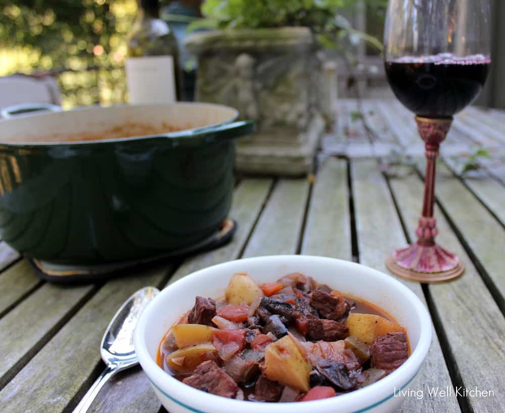 bowl of beef stew and pot of soup with glass of red wine on wooden table outside
