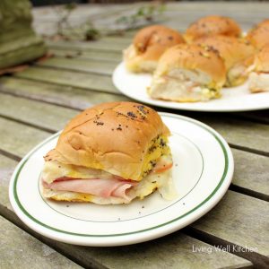 white plate with a ham and swiss slider with a plate of extra sliders in background on a wooden table.