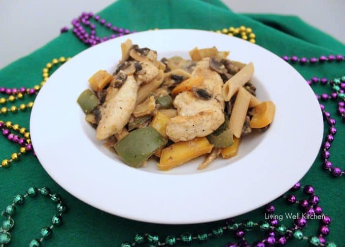white bowl of chicken, pasta, bell peppers, and mushrooms on green tablecloth with purple, green, and gold beads