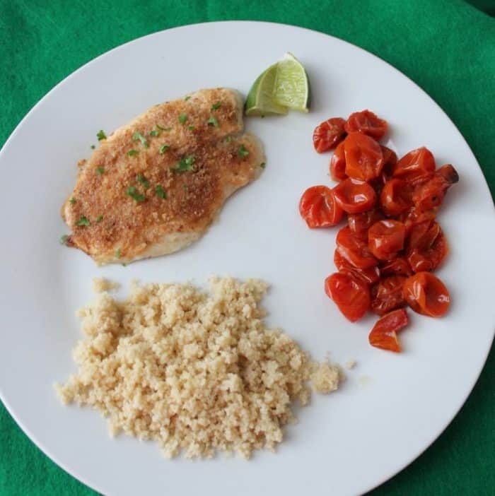 white plate of Easy Baked Fish, lime, roasted tomatoes and couscous on green table