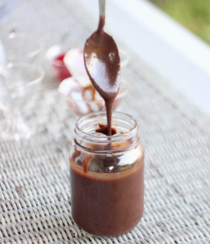spoon with chocolate sauce dripping off into a jar of chocolate sauce on a white table