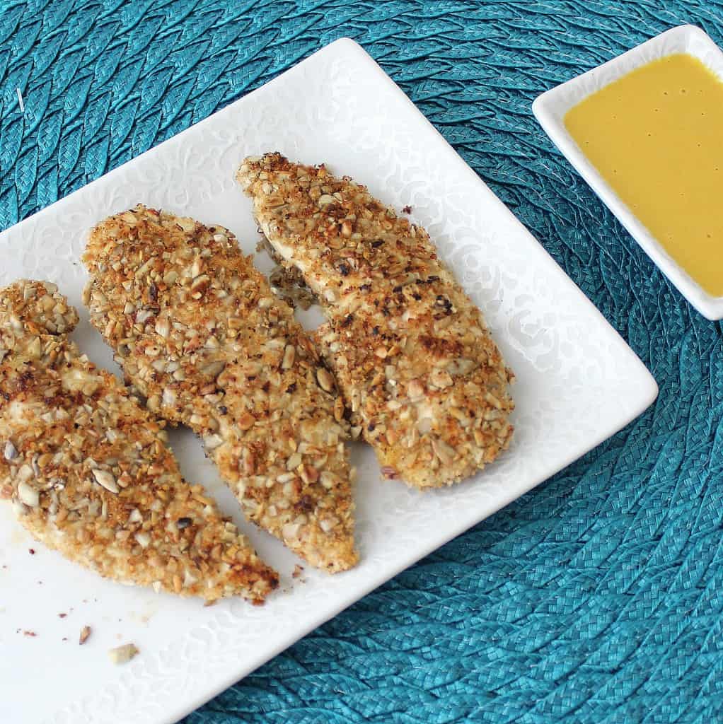 three sunflower seed chicken fingers on a white plate on a blue placemat with honey mustard