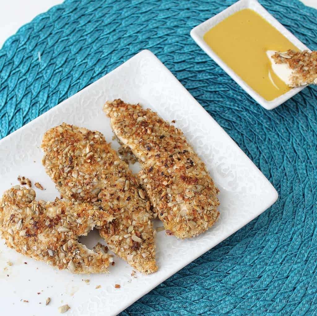 blue placemat with white plate of Sunflower Seed Crusted Chicken Fingers and chicken finger dipping into honey mustard sauce.