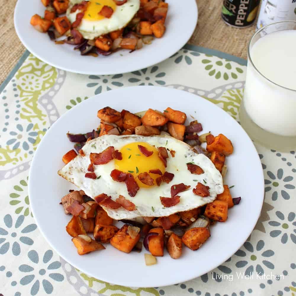 two white plates of sweet potato hash with fried egg and bacon on placemat with salt and pepper shakers and glass of milk