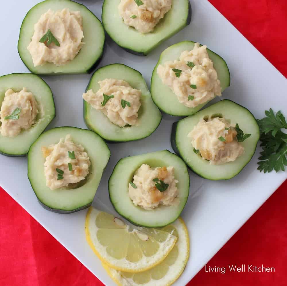 cucumber slices filled with roasted garlic hummus on a white plate with lemon slices.