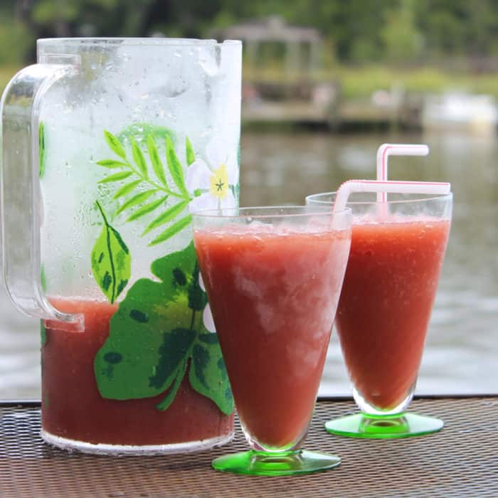 pitcher of and two glasses of Red Roosters with straws and water with a pier in the background