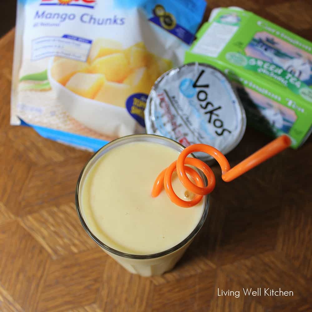 glass of mango green tea smoothie with an orange swirly straw next to a bag of frozen mango, a box of green tea, and a container of greek yogurt.