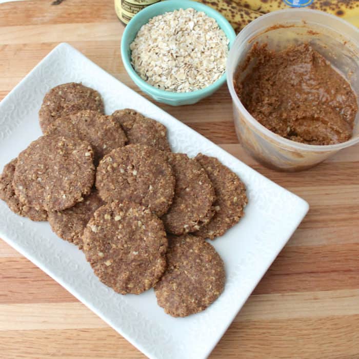 cookies on a white plate on a wooden cutting board with a bowl of oats and a container of almond butter