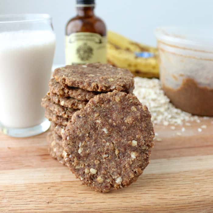 stack of almond butter banana cookies with one cookie facing the front on wooden cutting board with a glass of milk, banana, oats, vanilla extract, and almond butter container