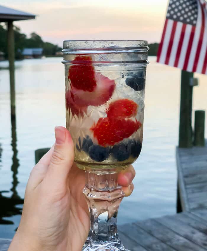 hand holding a glass made from a mason jar filled with red white and blue sangria and a river in the background