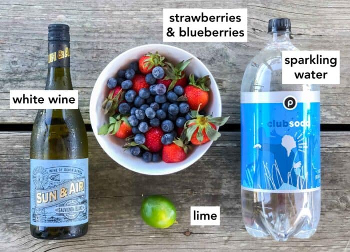 wooden table with white wine, bowl of strawberries and blueberries, one lime, and a bottle of club soda