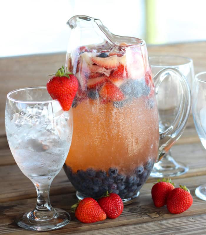 Glass full of ice with a pitcher of red white and blue Sangria with strawberries and blueberries