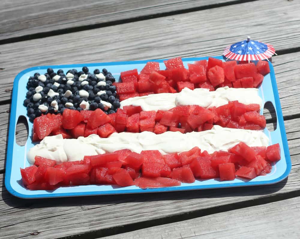 watermelon, blueberries, and whipped cream arranged like an American flag on a blue platter on wooden background with a cocktail umbrella with red white and blue design 
