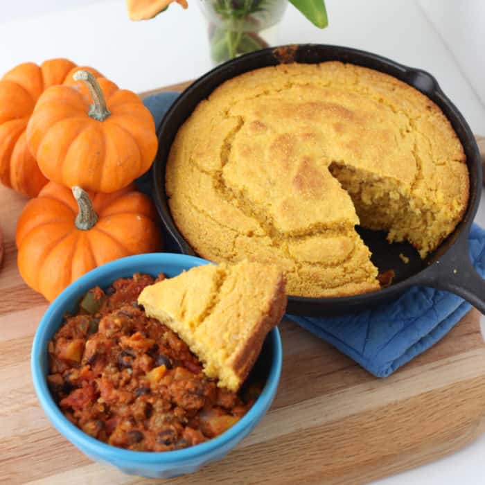 blue bowl of Pumpkin Chili with Pumpkin Cornbread in a cast iron skillet on wooden cutting board with three small pumpkins