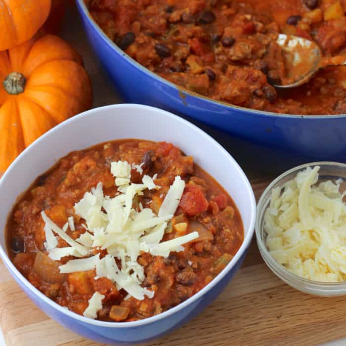 blue bowl of Pumpkin Chili with blue pot of chili and small bowl of cheese with small pumpkin