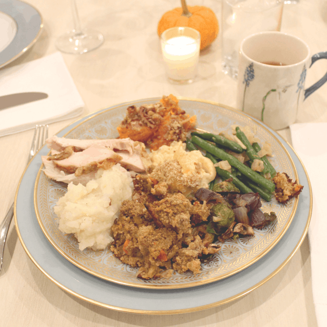 Thanksgiving plate with turkey, mashed potatoes, stuffing, roasted vegetables, green beans, squash casserole, sweet potato casserole on a white table with a mug of apple cider