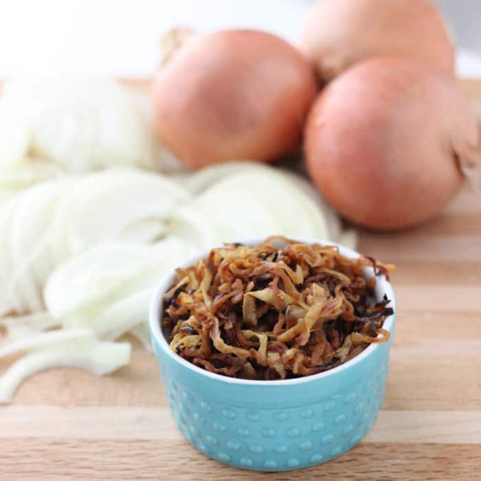 a blue bowl of Caramelized Onions with whole and sliced raw onions in the background on wooden cutting board