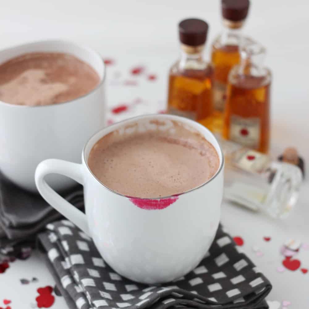 two white mugs one with a lipstick stain that have hot chocolate on white and grey towels, with heart confetti underneath and bourbon bottles in background