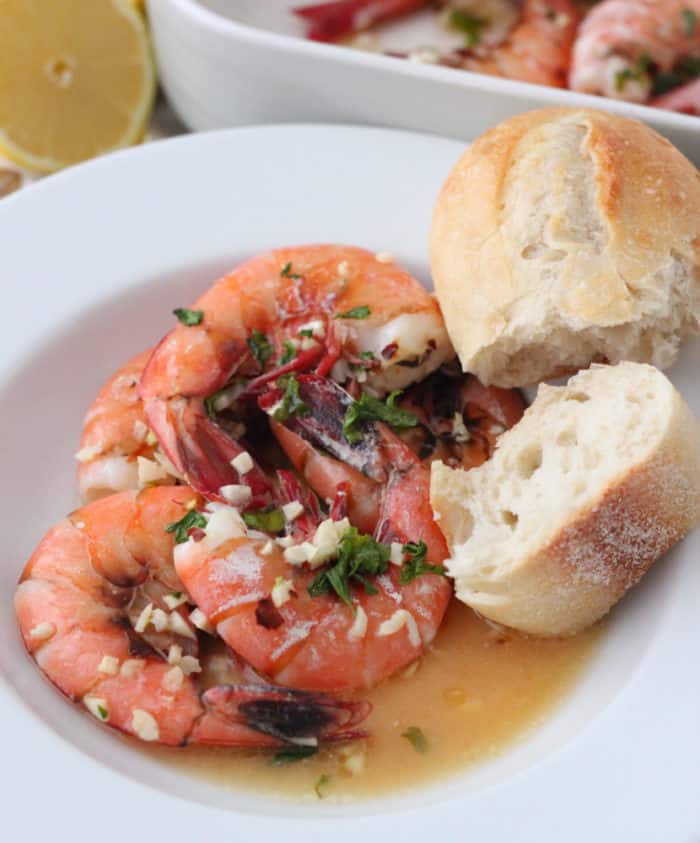 white bowl of shrimp covered in parsley, garlic and butter with french bread and lemon