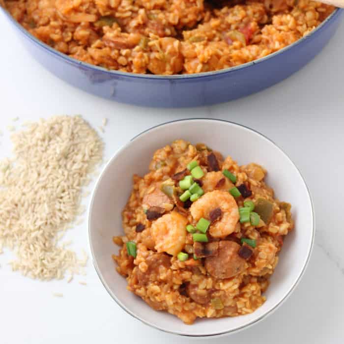 white bowl of shrimp and sausage Jambalaya with uncooked rice and blue pot of jambalaya