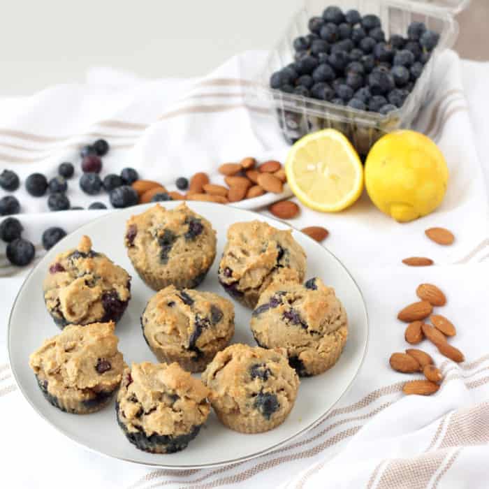 Blueberry Almond Muffins on white plate on white and tan dish towel with fresh blueberries, whole almonds and cut lemon