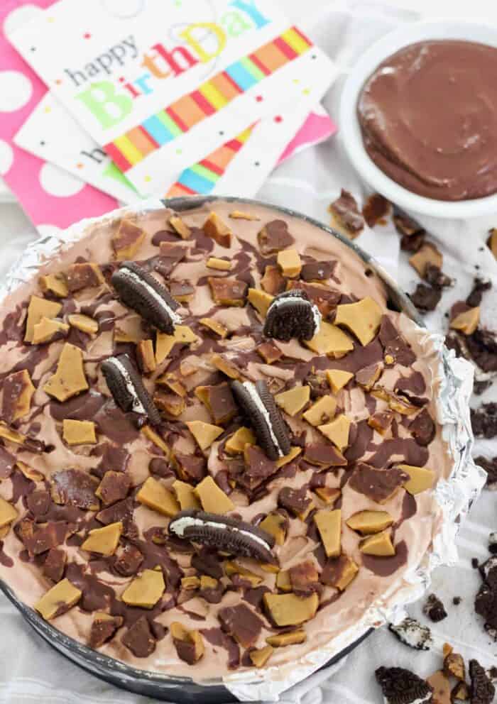 ice cream pie in a springform pan topped with oreo halves, homemade toffee and chocolate sauce. With a bowl of chocolate sauce, Happy Birthday napkins, and crushed cookies around pie