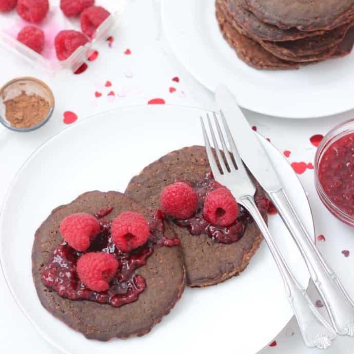 two Chocolate Protein Pancakes with raspberries and raspberry sauce on a white plate with fork and knife and valentines day confetti l;;;