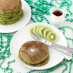 white plate with two green pancakes, knife and fork, sliced kiwi on a table with green beans, stack of pancakes, and a small container of syrup