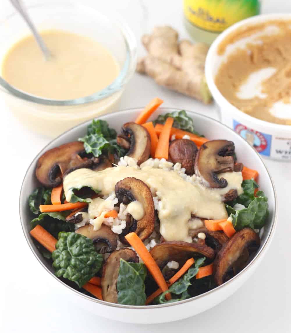 white bowl with kale, brown rice, carrots, and mushrooms topped with a miso sauce with miso, fresh ginger, and extra miso dressing in background