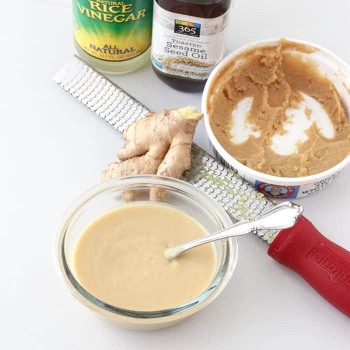 clear bowl of miso sauce with silver spoon next to a red microplane with fresh ginger, a container of miso paste, a bottle of sesame oil, and a bottle of rice vinegar