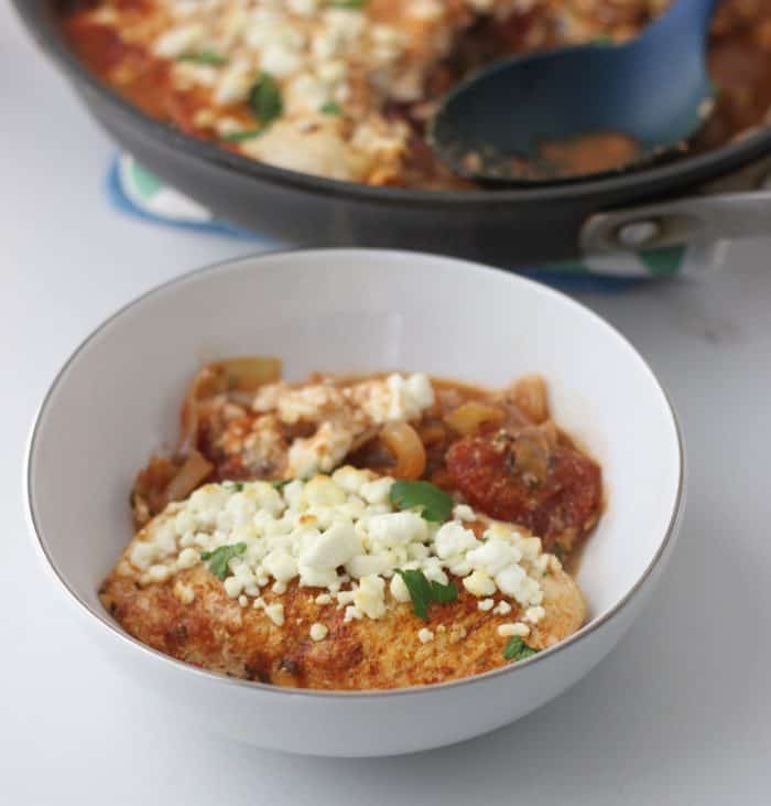 white bowl of chicken covered in onions, tomatoes and feta with a skillet in the background
