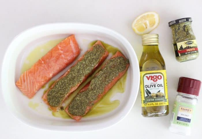 three salmon fillets in white baking dish covered with pesto, cut lemon, olive oil, pesto, salt