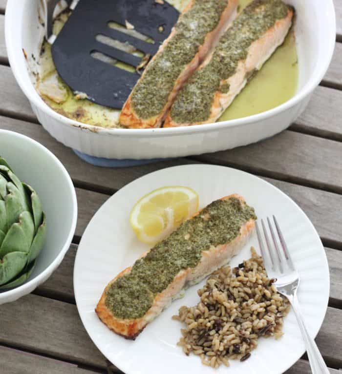 artichoke in white bowl, salmon, lemon slice, and wild rice on white plate, pesto covered salmon and spatula in white baking dish