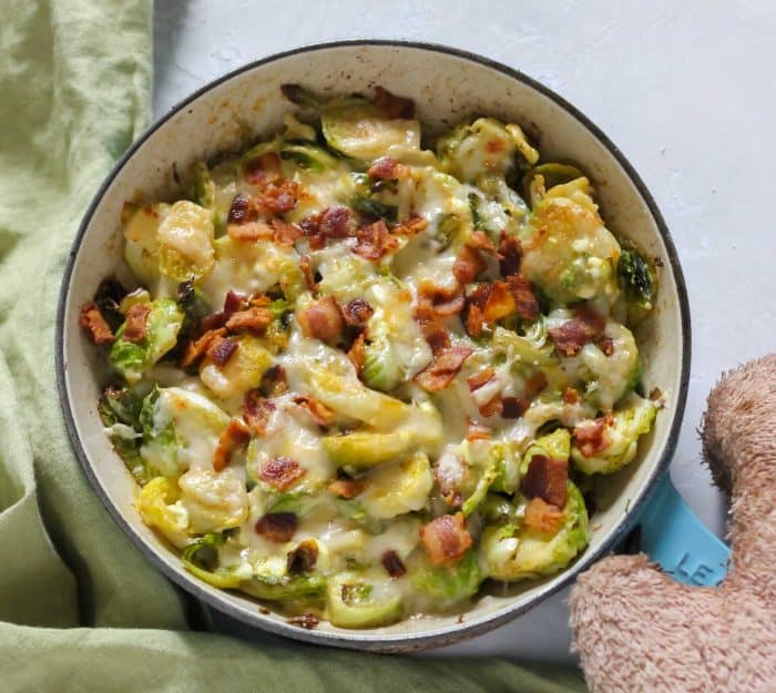 skillet with Brussels sprouts covered in cheese and bacon, oven mitt holding skillet and green napkin