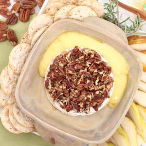 baking dish surrounded by crackers, pear slices, rosemary and pecans with baked brie topped with pecans and brie oozing out into baking dish