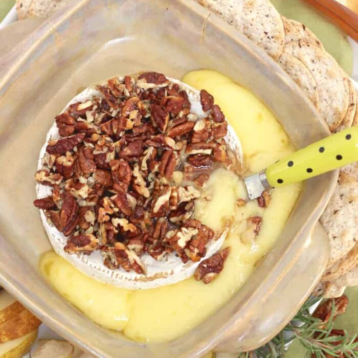 baking dish with baked brie covered in pecans with a small knife in brie and brie oozing out into baking dish surrounded by crackers