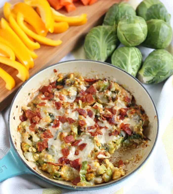 skillet of Cheesy Brussels Sprouts Dip with bell peppers on wooden cutting board and fresh Brussels sprouts