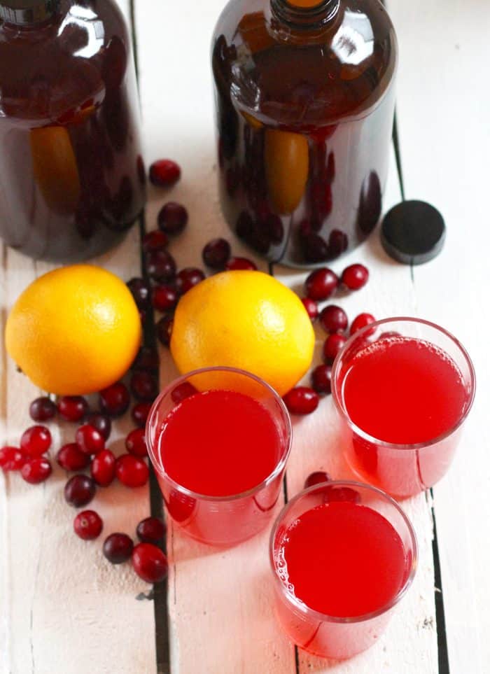 two large amber bottles with kombucha, two oranges, fresh cranberries, and three glasses of cranberry kombucha on a white wooden table