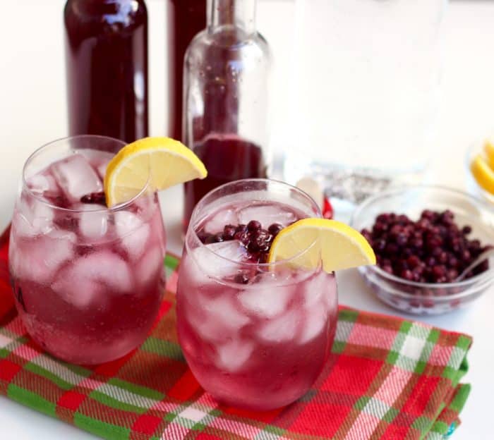 two purplish-blue cocktails made with blueberry lemon infused vodka and garnished with lemon slices, a bowl of frozen blueberries all sitting on top of a Christmas plaid napkin